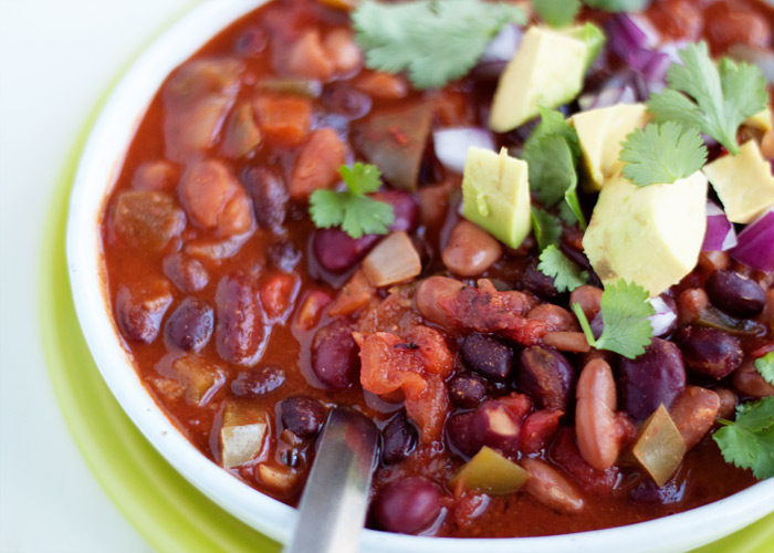 A bowl of 3-bean chili in a white bowl on a lime green plate with avocados, red onion, and cilantro on top. 