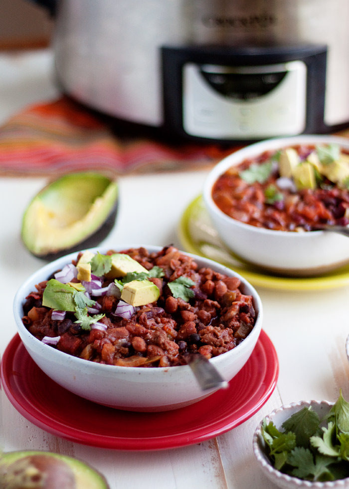 A bowl of 3-bean chili in a white bowl on a red plate with avocados, red onion, and cilantro on top and a Crock Pot in the background. 