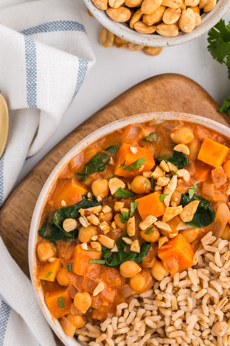 Top view of Slow Cooker Sweet Potato, Chickpea, & Peanut Stew in a white bowl with peanuts, salt and pepper, cilantro, and a Crock Pot in the background.