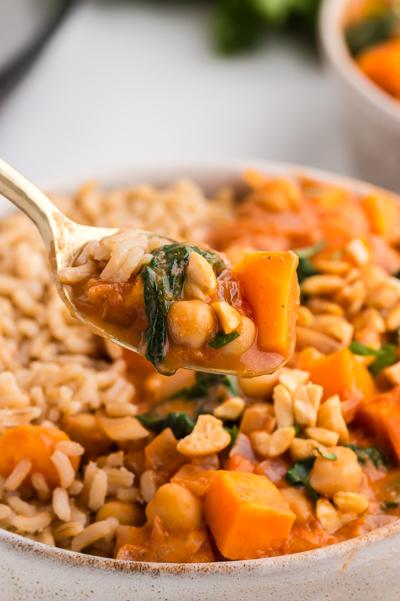 A gold spoon lifts out a bite of Slow Cooker Sweet Potato, Chickpea, & Peanut Stew from a white bowl.