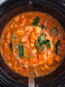 A ladleful of Slow Cooker Sweet Potato, Chickpea, & Peanut Stew is scooped out of a Crock Pot.