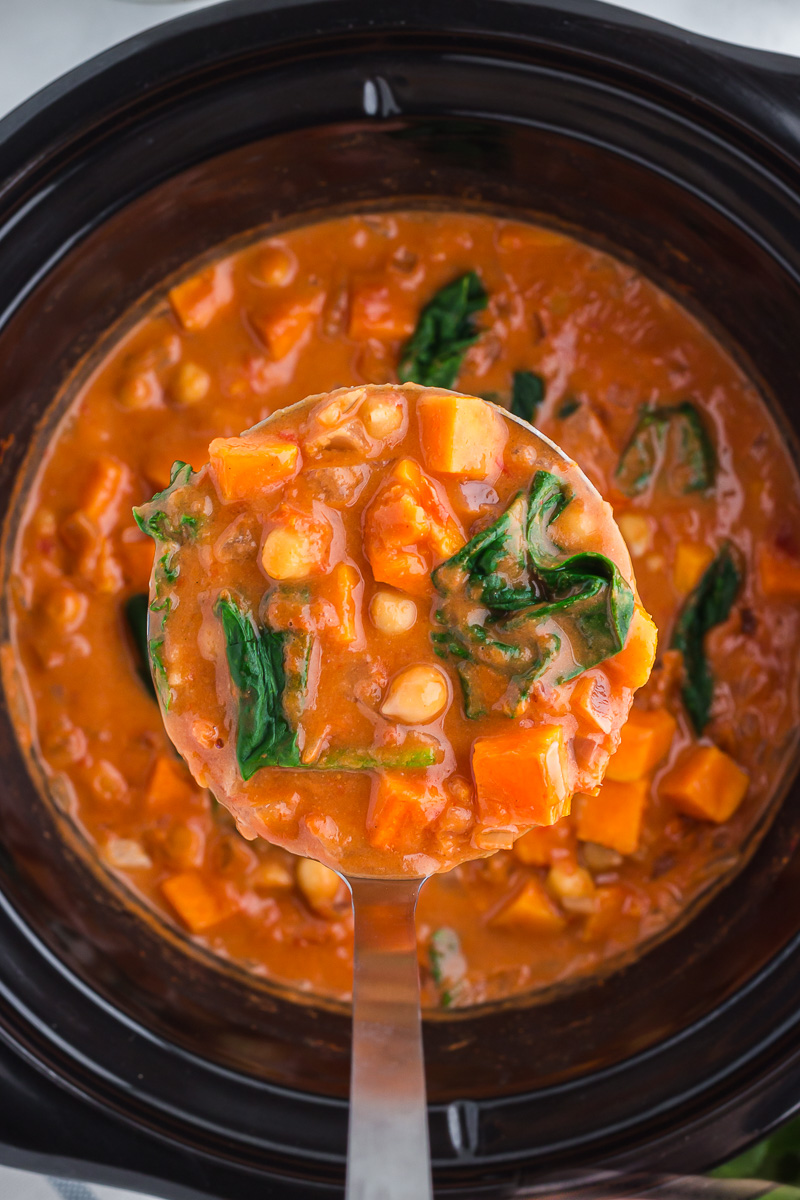 A ladleful of Slow Cooker Sweet Potato, Chickpea, & Peanut Stew is scooped out of a Crock Pot.