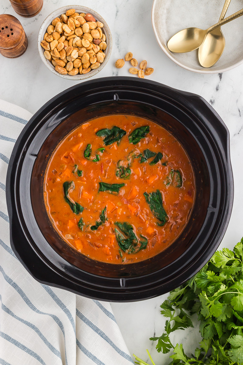 Slow Cooker Sweet Potato, Chickpea, & Peanut Stew in a black Crock Pot with peanuts and cilantro in the background.