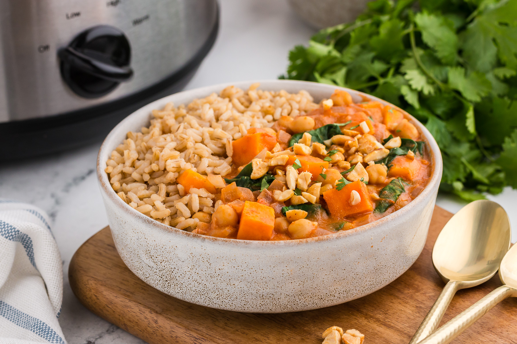 Slow Cooker Sweet Potato, Chickpea, & Peanut Stew in a white bowl with cilantro and a slow cooker in the background.