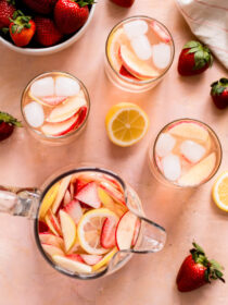 A top view of strawberry sangria in a pitcher, with glasses full of sangria and ice. The background is a peachy red.
