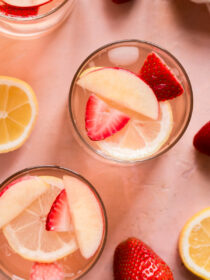 A top view of two glasses of sangria on a pink background with strawberries and lemons.
