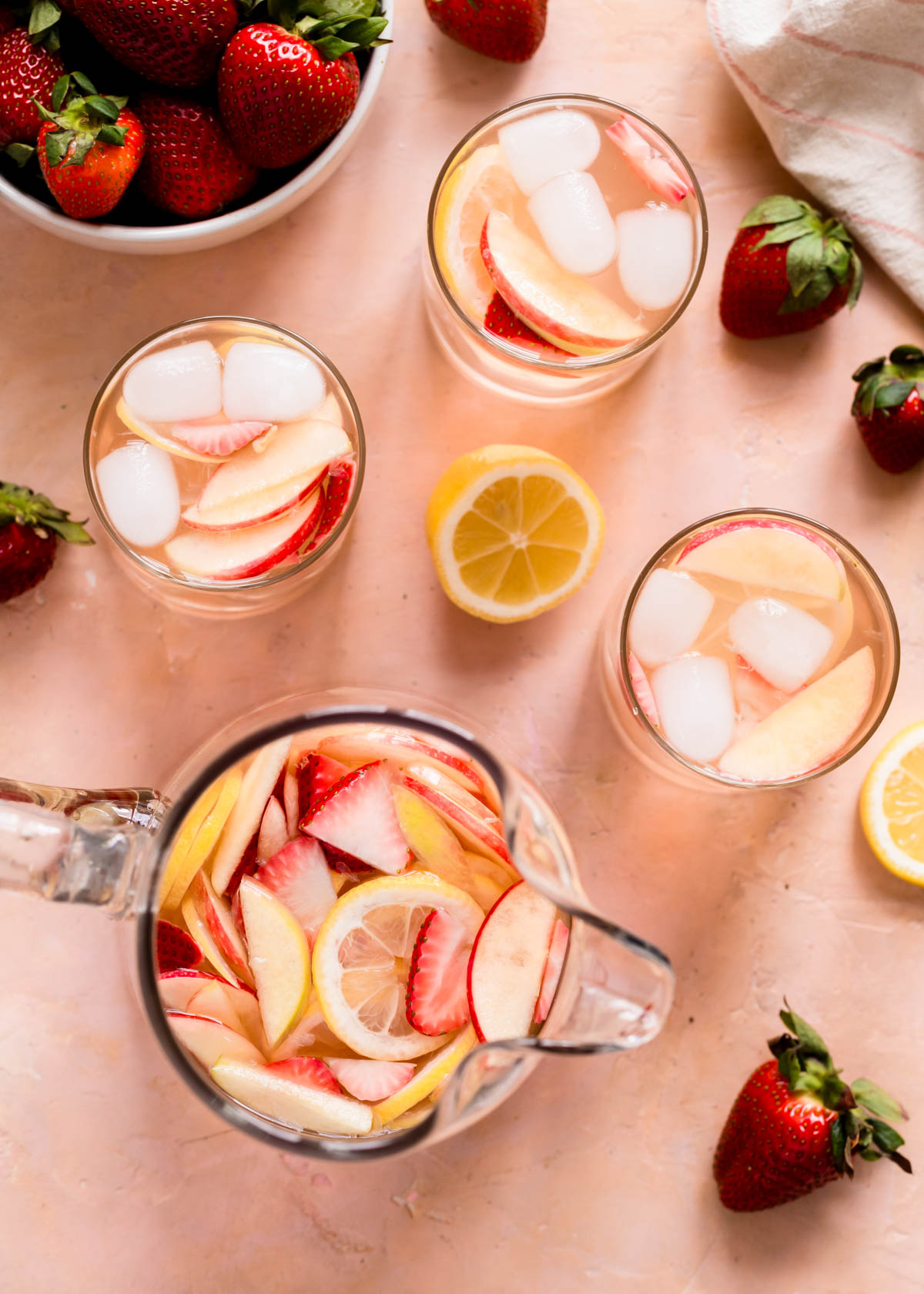 A top view of strawberry sangria in a pitcher, with glasses full of sangria and ice. The background is a peachy red.