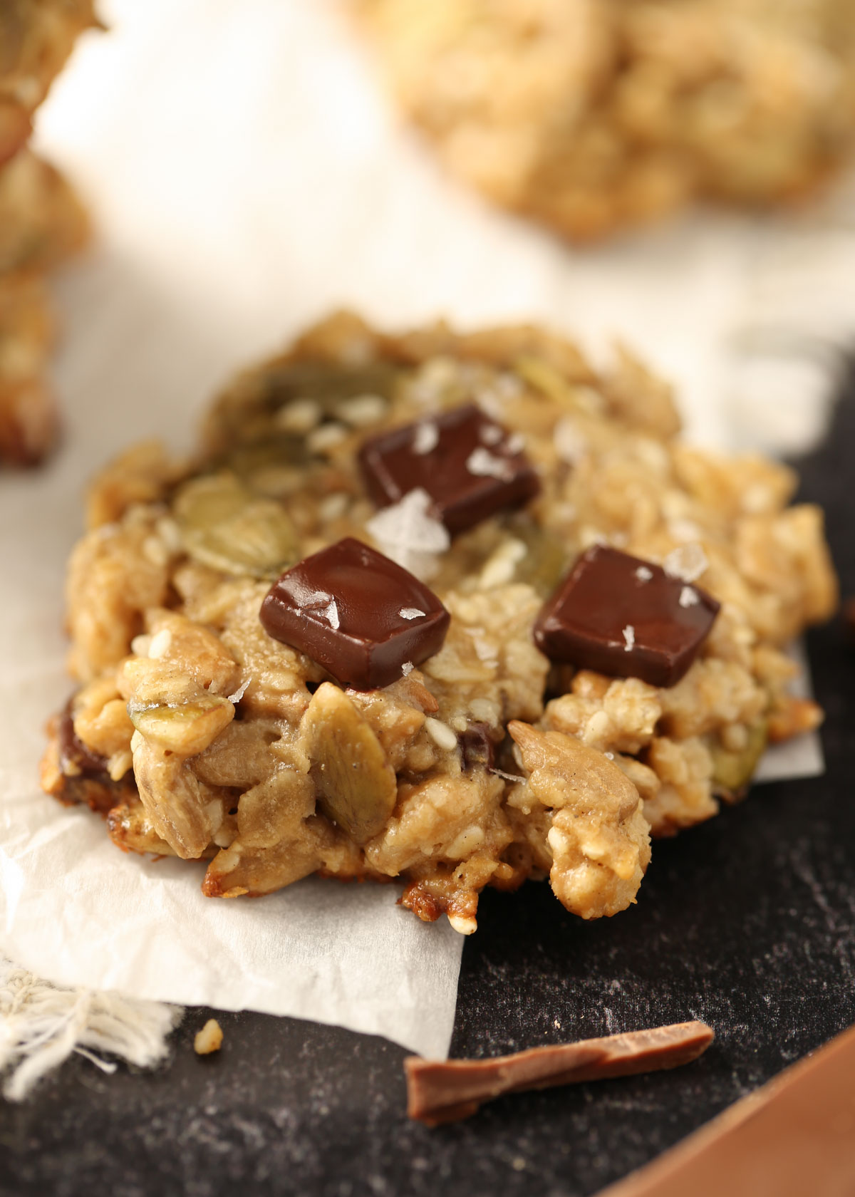 Close-up of a tahini seed cookie with chocolate chunks and flaky sea salt on a piece of parchment paper.