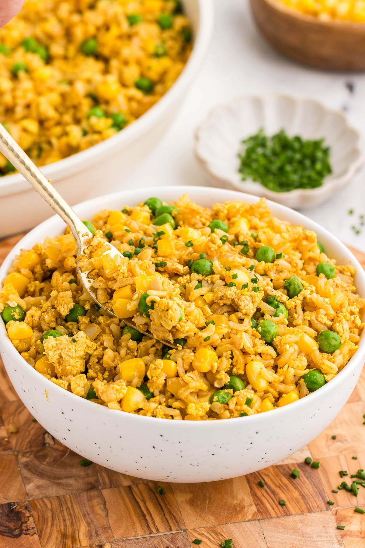 A white bowl full of tofu fried rice with a fork in it. A bowl of scallions rests in the background