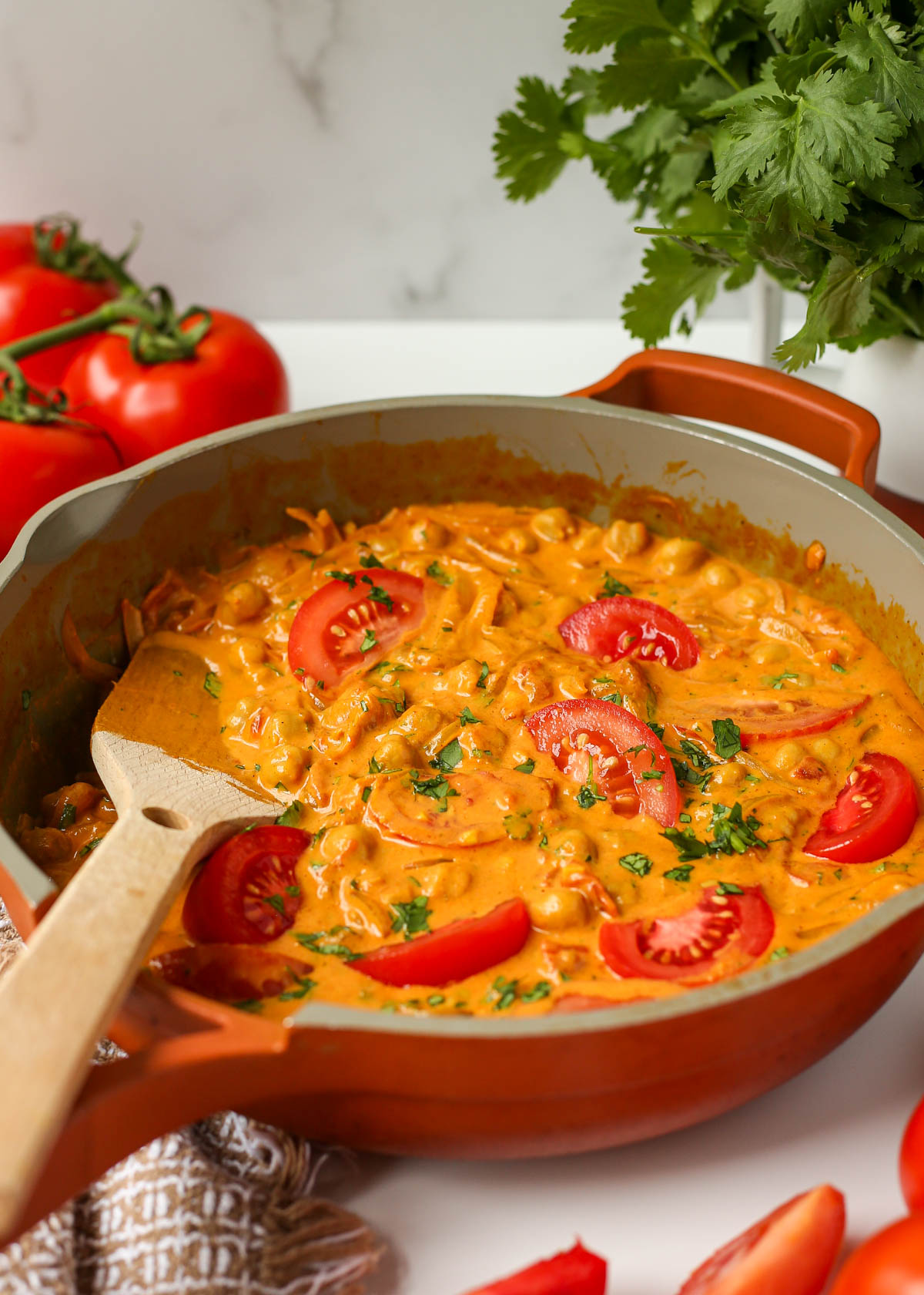 A pan with chickpea and tomato curry and fresh tomatoes and cilantro on top. Cilantro and tomatoes are in the background. 