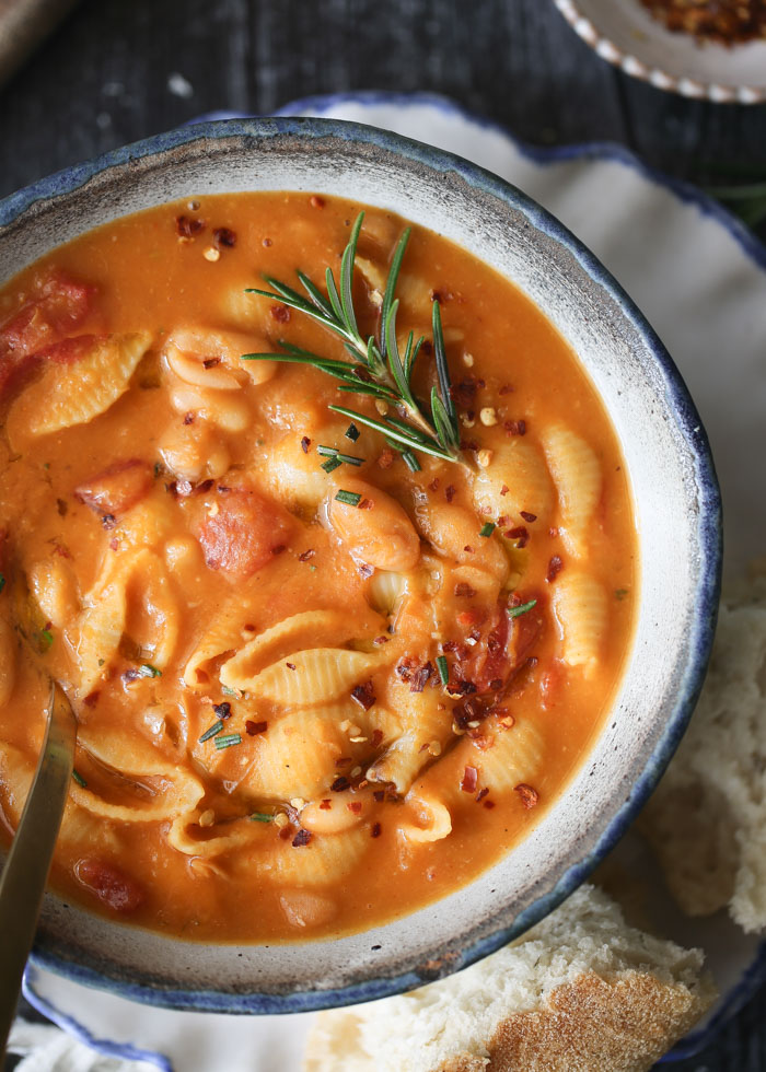 A top view of a rustic ceramic bowl of pasta e fagioli with a rosemary garnish.