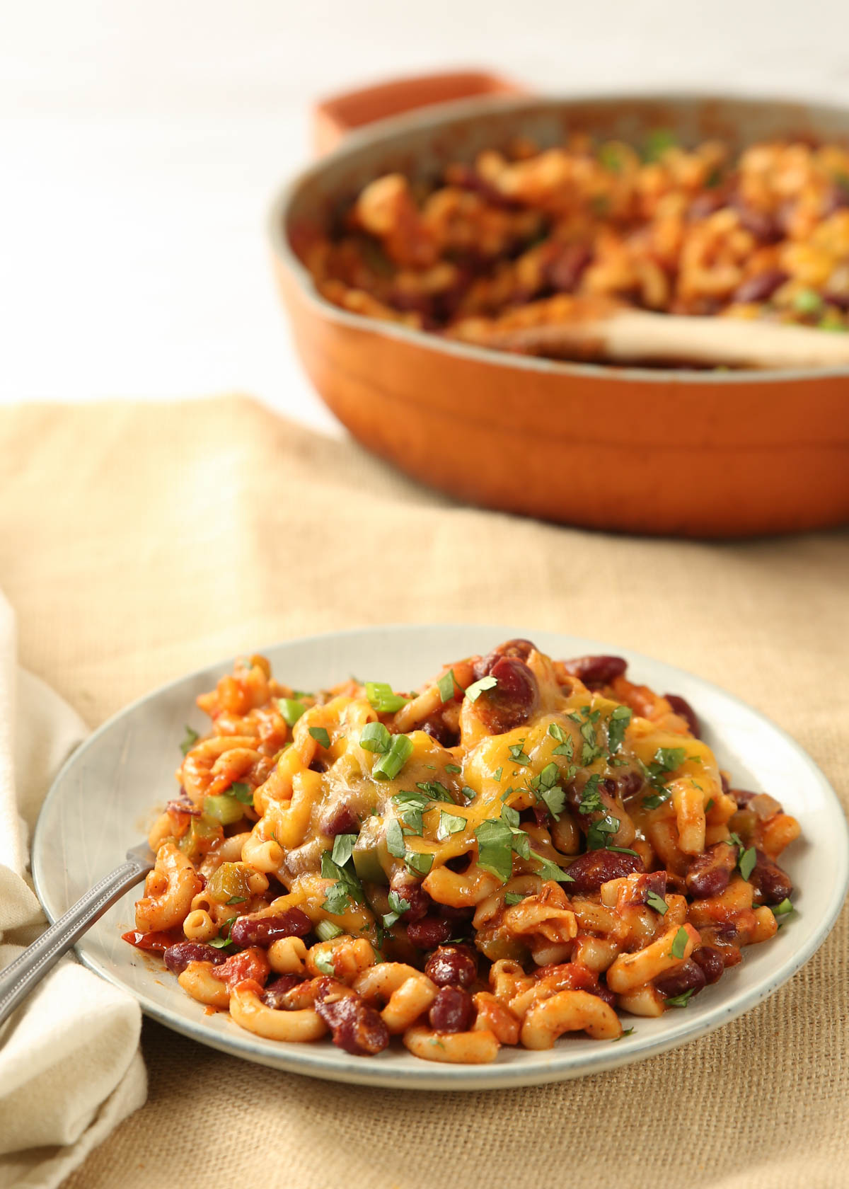 One-Pot Vegetarian Chili Mac on a plate with a fork, a beige background, and the pan of chili mac in the background.