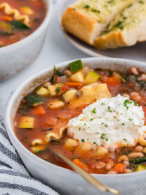 vegetable lasagna soup in a rustic ceramic bowl with a gray and white striped napkin and a gold spoon plus garlic bread in the background.