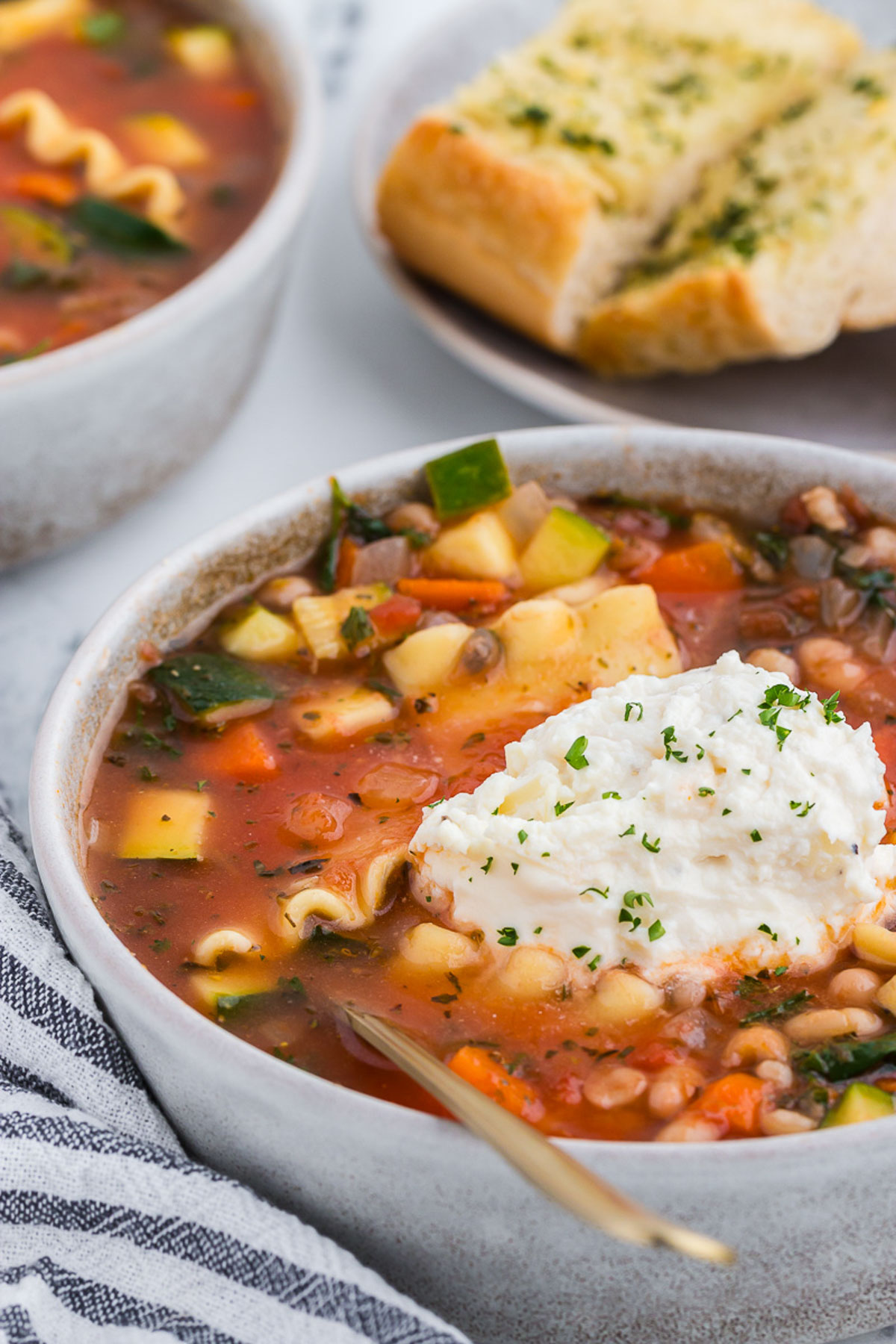 vegetable lasagna soup in a rustic ceramic bowl with a gray and white striped napkin and a gold spoon plus garlic bread in the background.