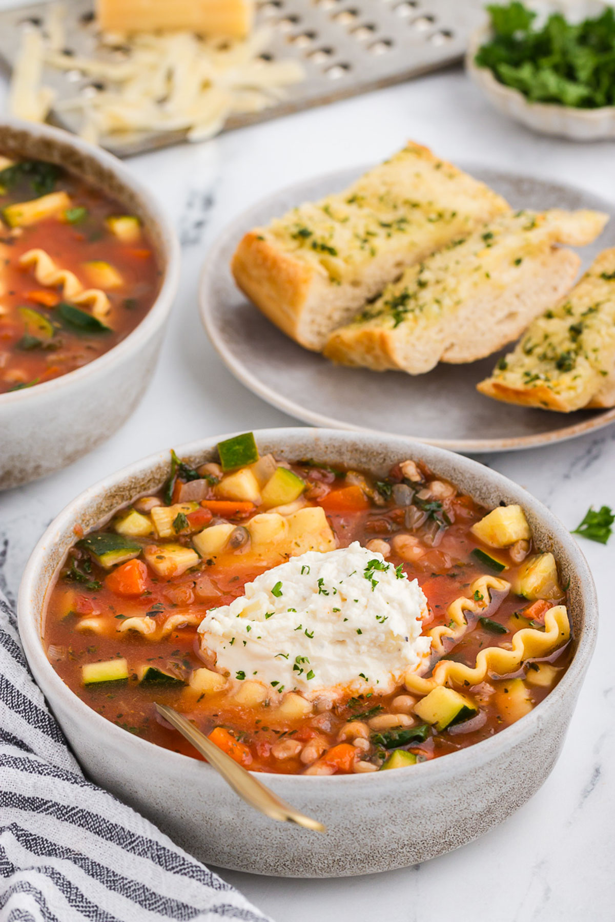 vegetable lasagna soup in a rustic ceramic bowl with a gray and white striped napkin and a gold spoon plus garlic bread in the background.