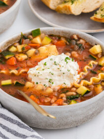 vegetable lasagna soup in a rustic ceramic bowl with a gray and white striped napkin and a gold spoon.