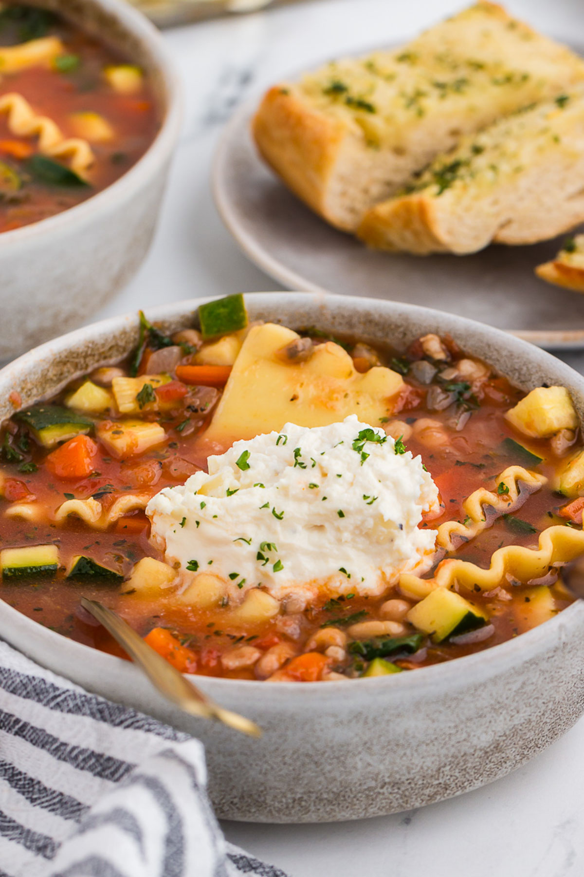 veggie lasagna soup in a rustic ceramic bowl with a gray and white striped napkin and a gold spoon.