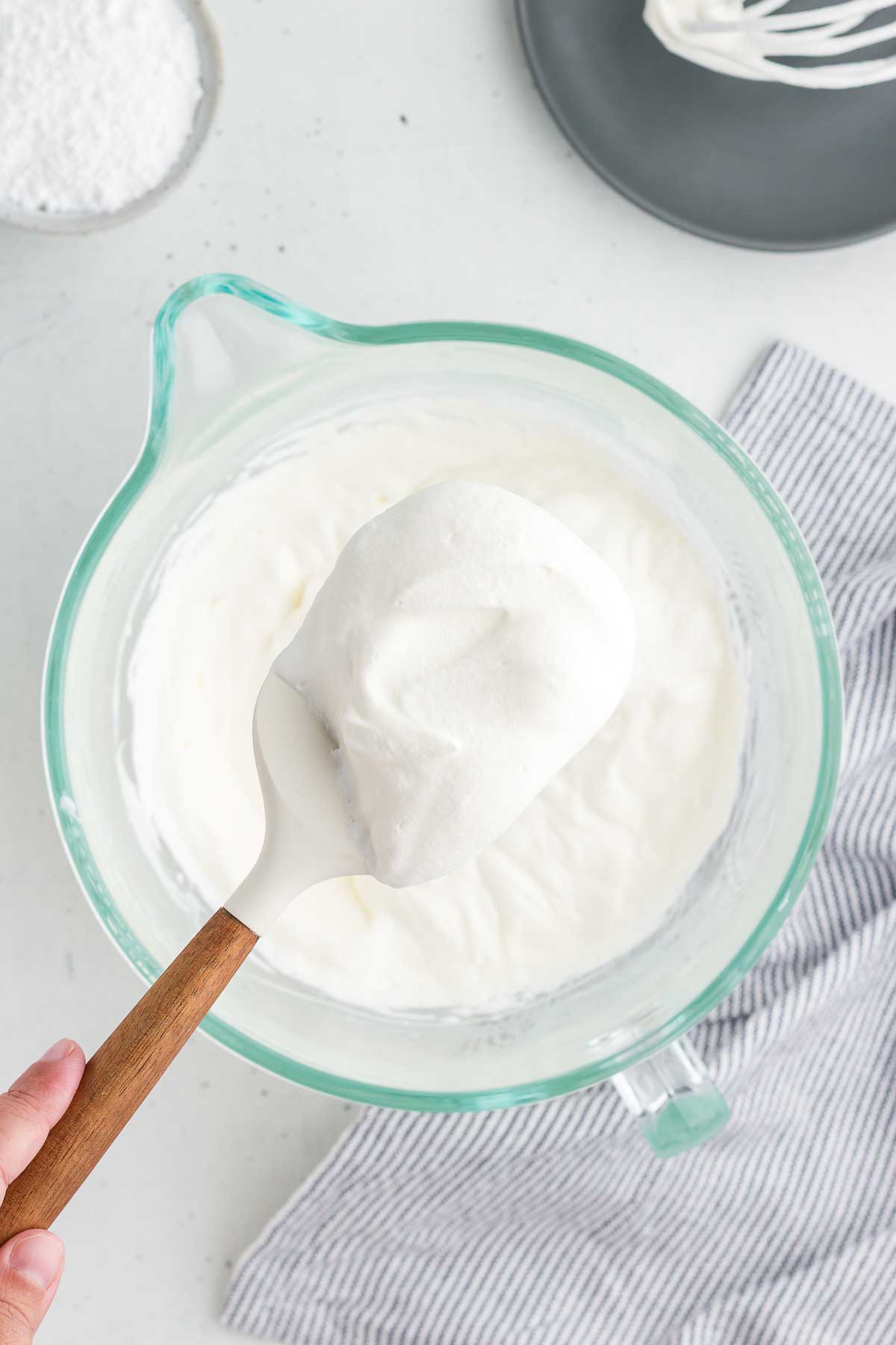 A spatula lifts out a scoop of rich creamy homemade whipped cream. A striped napkin is in the background.